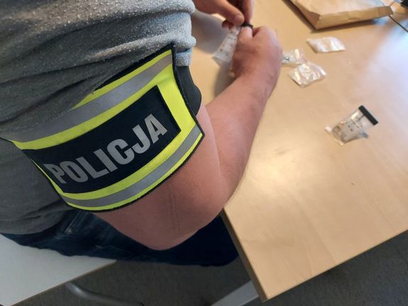 Policjant z opaską na przedramieniu z napisem policja siedzi przy biurku na którym leżą narkotyki w foliowych małych torebeczkach.