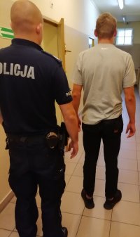 Zatrzymany mężczyzna stojący tyłem wraz z policjantem w pomieszczeniu dla osób zatrzymanych.