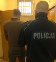 Zatrzymany mężczyzna oraz umundurowany policjant stojący tyłem w pomieszczeniu dla osób zatrzymanych.