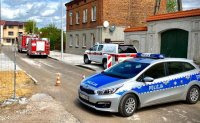 Radiowóz policyjny oznakowany, dwa radiowozy straży pożarnej oraz pogotowia gazowego na wąskiej ulicy w trakcie przebudowy.