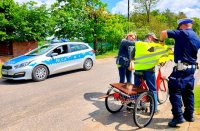 Policjant ruch drogowego oraz kobieta zakładają kamizelkę mężczyźnie stojącemu przy rowerze, dalej zaparkowany jest radiowóz policyjny oznakowany.