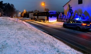 Autobus na poboczu, za nim bus, radiowóz z włączonymi światłami, domy w tle i śnieg.
