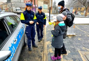Policjantki w mundurach, radiowóz i kobieta z dzieckiem otrzymująca odblask i kamizelkę.