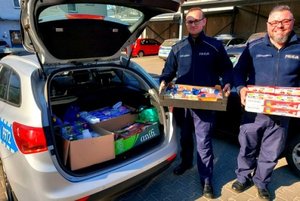 Policjanci w mundurach stoją przy radiowozie trzymając w rekach pudełka z produktami, bagażnik wypełniony produktami spożywczymi i chemicznymi.