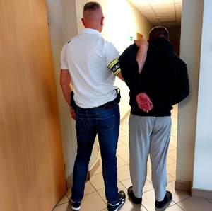 Policjant w ubraniu cywilnym z opaską na ręce z napisem policja na korytarzu trzyma za ramię zatrzymanego mężczyznę w kajdankach, obaj stoją tyłem.