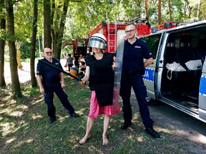 Kobieta ubrana w sprzęt policyjny kask, ochraniacz trzyma w ręce pałkę i tarczę oraz dwaj policjanci.