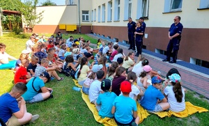 Policjanci oraz strażnicy miejscy oraz dzieci siedzące na trawie i słuchające mundurowych.