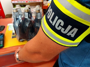 Ręka mężczyzny na której jest opaska z napisem policja i na ladzie sklepowej stoi zgrzewka piwa.