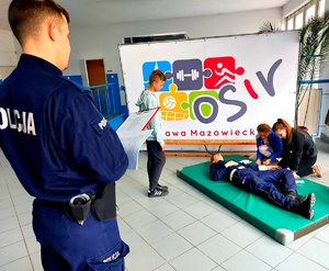 policjant oceniający dzieci które udzielają pierwszej pomocy przedmedycznej policjantce pozorującej osobę która zasłabła.