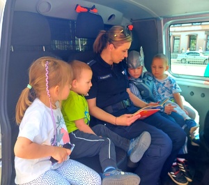 policjantka siedzi w radiowozie z dziećmi i czyta im książeczkę.