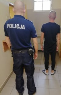 Zatrzymany mężczyzna stojący tyłem wraz z policjantem w pomieszczeniu dla osób zatrzymanych.