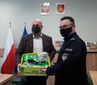 Wójt Gminy Rawa Mazowiecka przekazujący Zastępcy Komendanta rawskiej policji pudełko z elementami odblaskowymi.