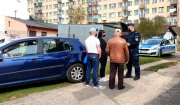 Policjant w mundurze oraz kobieta i dwóch mężczyzn stojących przy niebieskim samochodzie, w tle radiowóz oznakowany.