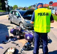 Policjant w kamizelce odblaskowej z napisem policja obok leży rozbity motor i szary samochód osobowy z wgniecionymi drzwiami.