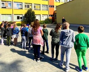 Dzieci stojące na placu szkolnym oraz policjant w mundurze prowadzący rozmowę z nimi.