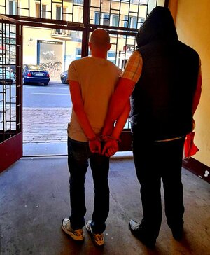 Dwóch mężczyzn stojących tyłem, jeden z nim ma kajdanki.