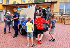 Policjant, radiowóz, dzieci wchodzące i wychodzące z radiowozu.