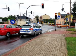 Radiowóz policyjny oraz inne auta na skrzyżowaniu z sygnalizacją świetlną.