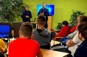 Policjant i policjantka w mundurze stoją przed młodzieżą i opowiadają o pracy w Policji, przed nimi tyłem do obiektywu siedzi młodzież.