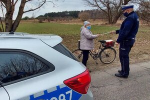 Policjant w mundurze dający odblask kobiecie stojącej przy rowerze i radiowóz oznakowany.