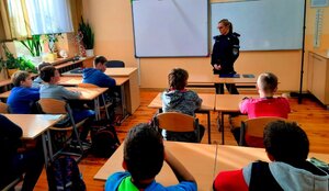 Policjantka w mundurze rozmawiająca z dziećmi siedzącymi w ławkach.