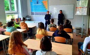 Policjantki w mundurach oraz młodzież siedząca w ławkach.