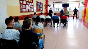 Policjanci, ksiądz i dzieci siędzące na krzesełkach na holu szkoły.