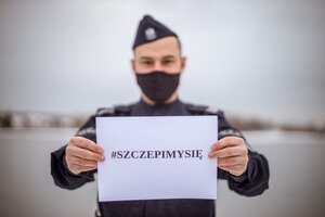 policjant w mundurze i w masce trzymający kartkę z napisem szczepimy się