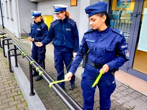 Trzy policjantki w mundurach wieszają odblaski na barierce przed komendą.