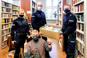 Dwóch policjantów trzymających pudełko z książkami, policjantka i kucający dyrektor biblioteki na tle regałów z książek.