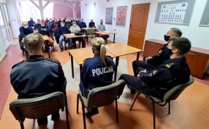 Policjantka i strażacy w mundurach siedzący na krzesłach przed stolikiem a przed nimi siedzą na krzesłach mężczyźni w maseczkach.