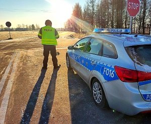 Policjant stojący przy radiowozie oznakowanym w umundurowaniu i kamizelce odblaskowej.