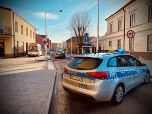 Radiowóz policyjny stojący w poprzek drogi, po lewej stronie karetka pogotowia na chodniku oraz na drodze samochód osobowy.