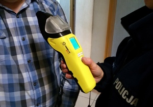 Urządzenie do badania stanu trzeźwości trzymane w rekach policjanta i kierowane do mężczyzny w koszuli w kratę.