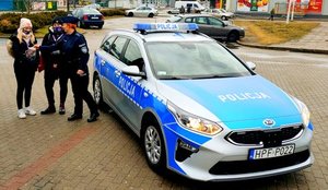 Policjantka wrzucająca pieniądze do puszki na Wielka Orkiestrę Świątecznej Pomocy, dwie wolontariuszki i radiowóz policyjny.