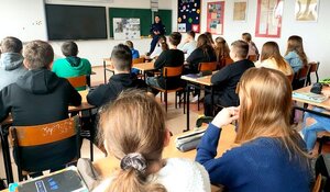 Policjantka w mundurze stojąca przed klasą, w ławkach siedzą uczniowie.