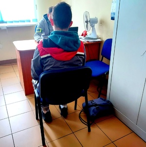 Mężczyzna siedzący na krześle tyłem, przy biurku policjant w cywilnym ubraniu.