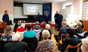 Policjanci w mundurach stoją przed seniorami siedzącymi na krzesłach w sali.