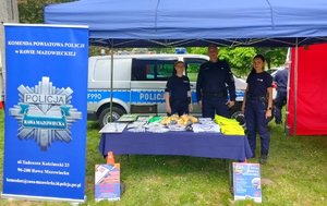 Policjant i dwie policjantki stojące pod namiotem przed nimi stół z przygotowanymi elementami promującymi bezpieczeństwo.