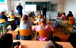 Policjant i policjantka stoją w klasie przed dziećmi, które siedzą w ławkach.