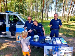 Dwóch policjantów i policjantka w mundurze obok radiowozu przy stoliku z materiałami promującymi bezpieczeństwo nad woda i dziecko.