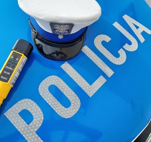 Czapka policjanta ruchu drogowego ma masce radiowozu z napisem policja oraz urządzenie do badania trzeźwości.