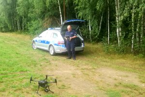 Policjant w mundurze przy radiowozie i dron na ziemi, w oddali las.