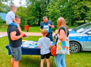 Policjant w mundurze na pikniku na stoliku ulotki i dorośli z dziećmi obok radiowóz.
