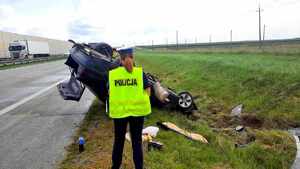 Policjantka w kamizelce z napisem policja stoi obok auta leżącego na dachu.