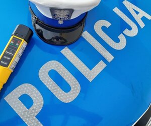 Napis policja na pokrywie silnika radiowozu, czapka policjanta ruchu drogowego oraz urządzenie do mierzenia trzeźwości.