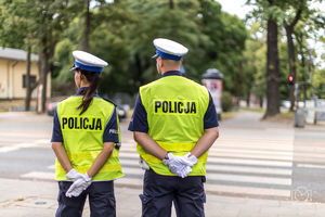 Policjant i policjantka w mundurze stoją tyłem przy przejściu dla pieszych.
