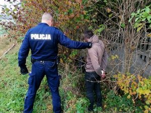 Policjant w mundurze trzyma zatrzymanego mężczyznę, który ma kajdanki na rękach w tle w krzakach schowane skradzione elektronarzędzia.