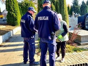 Policjanci w mundurach rozmawiają na cmentarzu z osobami sprzątającymi groby.
