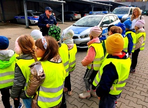 Dzieci w kamizelkach przysłuchują się rozmowie policjanta w mundurze, dalej radiowozy i nauczycielka.